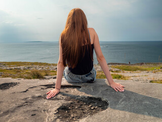 A slim woman wearing a black tank top sits on a rock and looking at Aran Island, Burren area of...