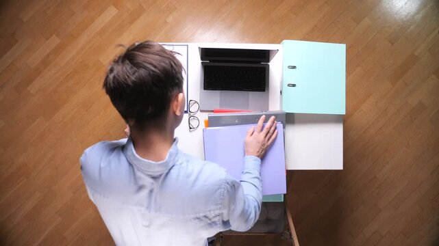 Employee carrying a stack of colorful ring binders and documents to an office desk with a laptop, clipboard, eyeglasses, and an empty chair, indicating workload and busy office environment