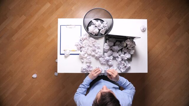 Frustrated person sitting at a desk and throwing many crumpled pieces of paper onto a laptop and into a wastebasket, symbolizing stress and creative failure