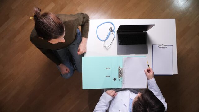 Doctor holding a medical file, discussing health information with a patient during a consultation, with a laptop, stethoscope, and clipboard visible on the white desk, captured from a high angle
