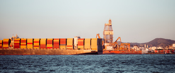 Cargo container ship passing an offshore oil rig near coastal port city at sunset, stacked containers on deck, calm sea and skyline, global trade and energy industry