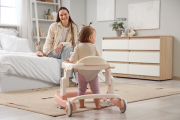 Cute little baby girl using walker with her mother in bedroom, back view
