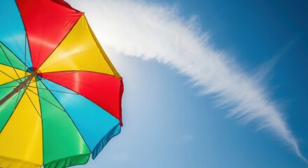 A vibrant, multi-colored beach umbrella stands out against a clear blue sky with delicate, wispy clouds.