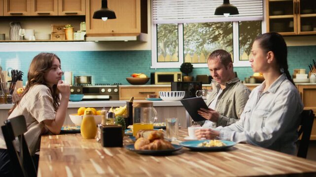 Working dad on tablet ignoring daughter and family at breakfast table, absorbed disconnected man distracted by his remote work. Inattentive parent distant with his girl and wife. Camera A.