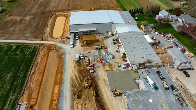 Workers operate machinery at a construction site in a rural area. Buildings are being developed and land is being prepared. Trucks are parked while work continues on different sections.