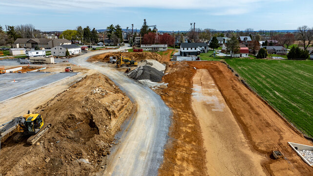 Workers use machinery at a construction site where dirt is moved and land is prepared for new buildings. Nearby homes and open fields show the suburban landscape changing over time.