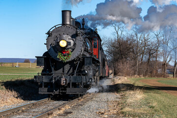 A steam locomotive pulls a set of cars along railway tracks surrounded by open fields. Smoke rises...