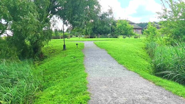 A person walks along a gravel path in a green park setting. Trees and a wooden fence border the path. The sky is cloudy and the grass is lush.