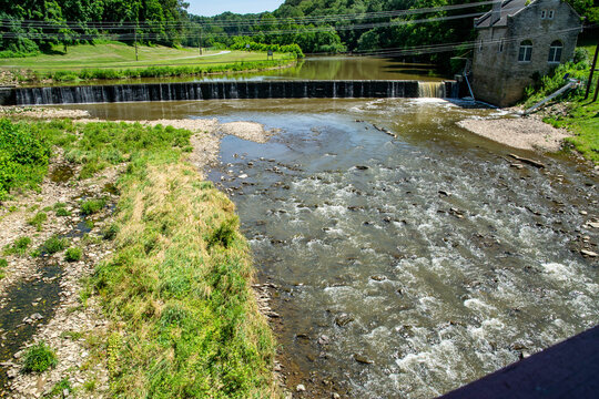 Water moves over a small dam creating ripples in the stream below. A stone building stands near the water in a green landscape. Trees and grass surround the area under a clear sky.