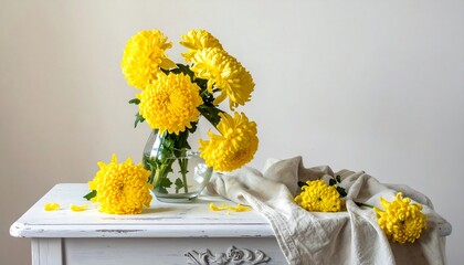 Bright yellow pompom chrysanthemums in a glass vase on a white table with a linen cloth