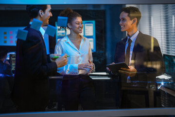 Diverse corporate team brainstorming ideas on glass wall. Businessman writing project plan with marker while female colleague analyzes sticky notes in modern dark office at night.