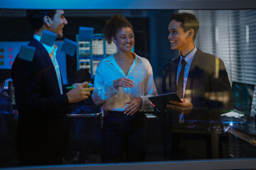 Diverse corporate team brainstorming ideas on glass wall. Businessman writing project plan with marker while female colleague analyzes sticky notes in modern dark office at night.