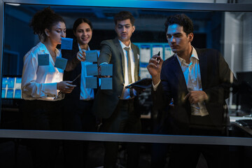 Agile tech startup team conducting scrum meeting on glass wall. Developers and managers mapping software workflow with sticky notes in modern blue-lit office at night.