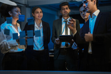 Agile tech startup team conducting scrum meeting on glass wall. Developers and managers mapping software workflow with sticky notes in modern blue-lit office at night.