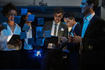Corporate marketing team planning business growth strategy. Executives analyzing financial goals and market trends using sticky notes on glass wall in modern office.