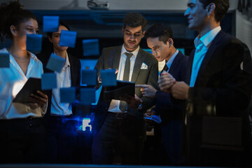 Corporate marketing team planning business growth strategy. Executives analyzing financial goals and market trends using sticky notes on glass wall in modern office.