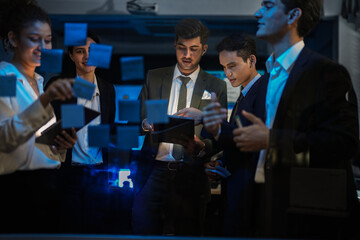 Obraz premium Diverse business team brainstorming project ideas on glass wall. Professional group discussing strategy with sticky notes in modern dark office during late night meeting.