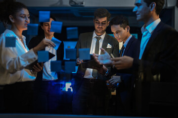 Obraz premium Diverse business team brainstorming project ideas on glass wall. Professional group discussing strategy with sticky notes in modern dark office during late night meeting.