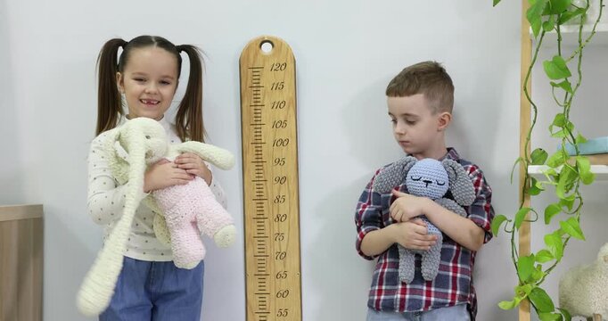 Little boy and girl measuring their height with chart near light wall indoors. Camera moving right