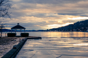 Winter sunset over Greenwood Lake, NY, with a quiet gazebo, fractured ice along the shoreline, and golden light reflecting across the water, creating a peaceful lakeside scene.