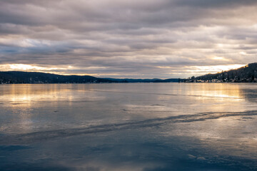 Calm winter view across Greenwood Lake, NY, with smooth ice and golden reflections beneath heavy...
