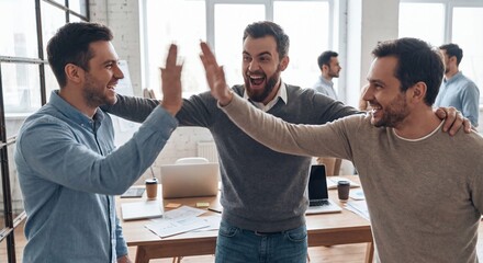 Three colleagues high fiving in a modern office with laptops and natural light teamwork and celebration with success and partnership and cooperation