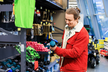 Smiling man in red jacket holding blue dumbbell while shopping for sports equipment in a retail store