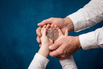Adult hands gently holding child hands with a silver metal heart shape against a blue background