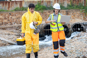 Environmental Engineers in PPE Suits Inspecting Wastewater Discharge from Industrial Pipe, Specialists Using Laptop for Water Quality Analysis and Pollution Monitoring at Drainage Site
