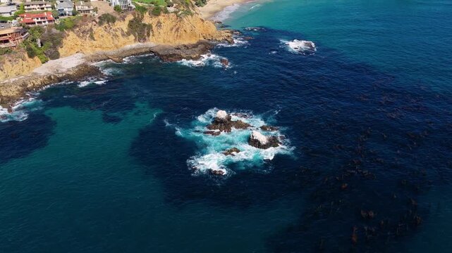 Aerial view of a coastal residential area with sandy coves and rocky cliffs, turquoise ocean water, and hillside homes stretching along the shoreline under clear sunny weather.