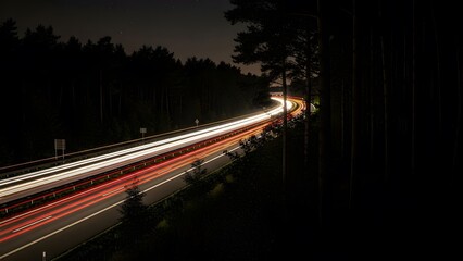 Long exposure shot of a highway at night, showing bright red and white light trails from cars passing through a dense forest under a dark sky with faint stars.