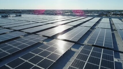 Array of Solar Panels - An array of solar panels are arranged on a flat rooftop, glistening in the sunlight. In the background is a cityscape with buildings. - Powered by Adobe