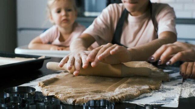 Close-up hands of teen girl rolls ginger cookie dough on a floured kitchen surface while a mother and her cute baby sister continue their warm homemade baking routine, family bonding concept