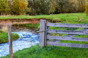 Wooden gate by Geleenbeek River with water flowing in Absbroekbos conservation area, greenish wild grass and trees in background, winter day in Munstergeleen, Limburg, Netherlands