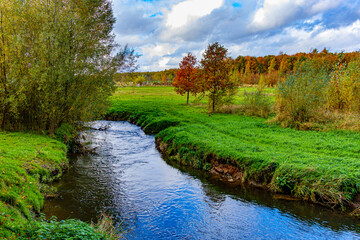 Plain of Absbroekbos conservation area with Geleenbeek river in foreground, shrubs and trees with lush green, yellow and red foliage in background, day in Munstergeleen, Limburg, Netherlands