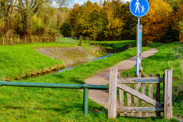 Post with pedestrian path sign at entrance to Absbroekbos conservation area, Geleenbeek river and trees with lush green, yellow and red foliage in background, Munstergeleen, Limburg, Netherlands