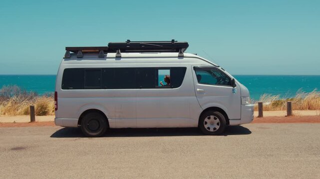 Young adult woman sits inside her van enjoying a snack while parked near the ocean in sunny weather.