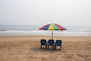 Quiet Puri beach with colorful umbrella and empty chairs facing gentle waves, soft sand, calm horizon under hazy sky morning.
