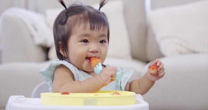 Asian toddler girl smiling happily while eating fruit at baby tray, concept of early learning, nutrition, family lifestyle, adorable childhood growth, self feeding, joyful home development moment
