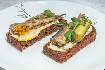 Two slices of dark rye bread topped with sprats, mustard seeds, lemon, and greens on a white plate.