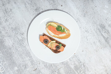 Top view of two delicious smoked salmon sandwiches with black olives and greens on a white plate over a textured gray background.