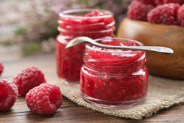 Homemade organic raspberry jam in small glass jars and a wooden bowl of fresh, healthy red raspberries on a rustic kitchen table.
