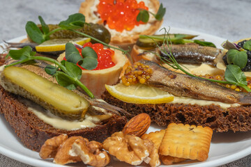 Close-up of assorted gourmet appetizers on bread with fish, pickles, nuts, and red caviar, on a white plate.