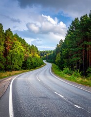 Fototapeta premium Winding asphalt road cuts through a vibrant forest under a partly cloudy sky. The road curves into the distance