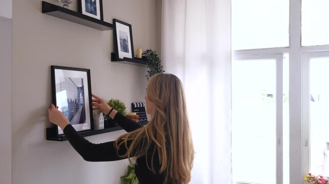 Woman adjusting a picture frame on the shelf in a modern home interior. Simple home decoration process, interior styling, lifestyle and everyday domestic activity