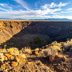 Arid landscape view of a large, steep-walled caldera under a blue sky with wispy clouds and distant hills
