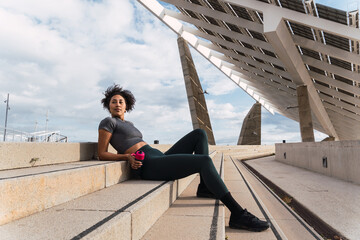Active young woman taking a break on stairs next to solar panels, holding a water bottle