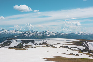 Scenic alpine landscape with sunlit snowy field on stony hill with view to big mountain range with forest and snow far away under cloudy sky. Stone outcrops among snows in sunlight in high mountains. © Daniil