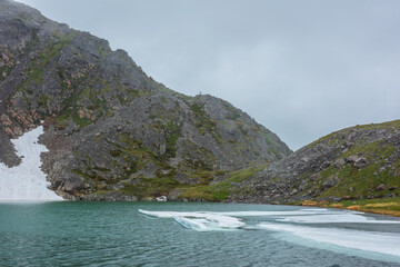 Ice floes float in alpine lake in rain. Ripples on turquoise water surface of beautiful lake against rocky pass under cloudy sky. Mountain lake among green hills under low clouds in rainy weather.