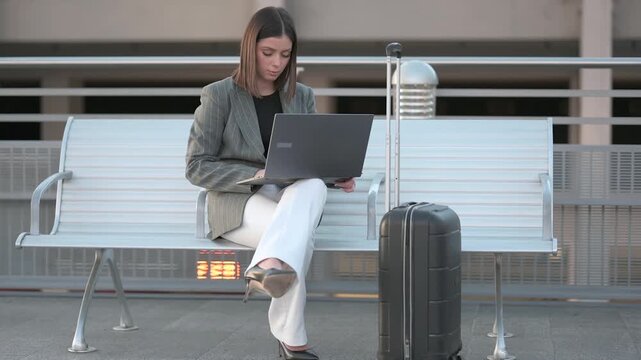 Confident female entrepreneur sitting on a bench with her luggage, typing on a laptop and checking emails while waiting for her transport at the airport or train station for a business trip. Slow moti